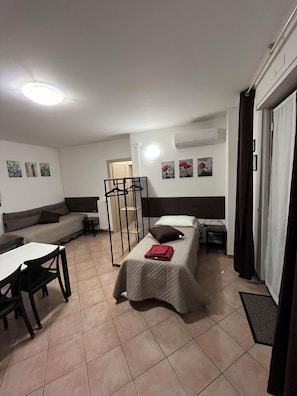 A compact hotel room with a bed, a sofa, a small table, and a chair. There is a window with curtains, an air conditioning unit, and a radiator. - Quadrifoglio affittacamere (Novi Ligure)
