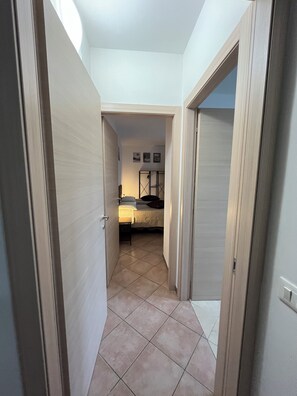 A narrow hallway leading to a bedroom with a bed, a nightstand, and a wall-mounted shelf. - Quadrifoglio affittacamere (Novi Ligure)