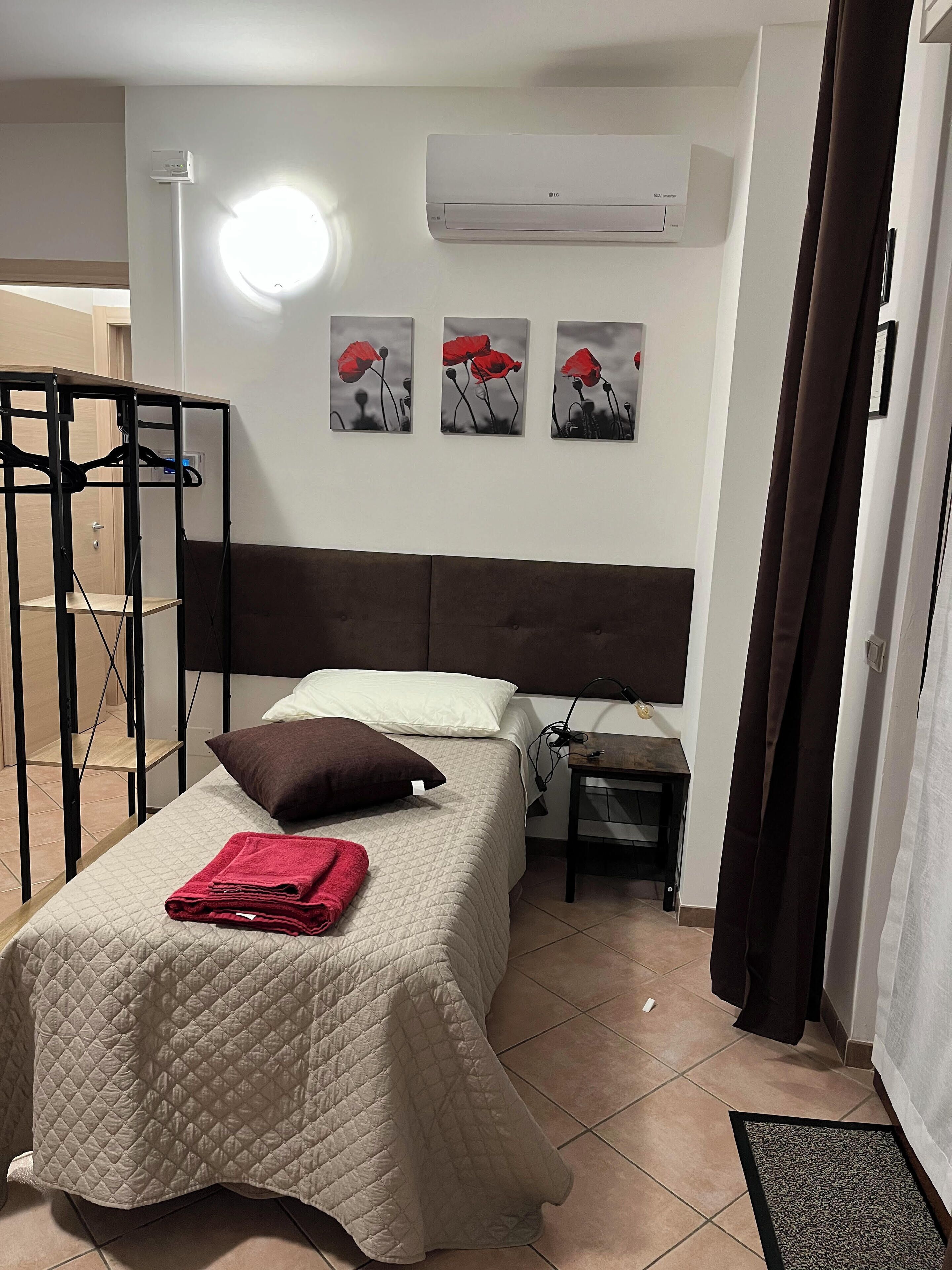 A hotel room with a bed, a bedside table, an air conditioning unit, and a wall-mounted artwork of red flowers.