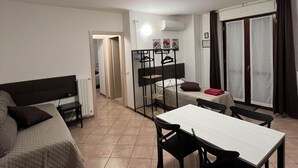 A compact room with a bed, a small table, and a chair. There is a window with curtains, a radiator, and a door leading to another room. - Quadrifoglio affittacamere (Novi Ligure)