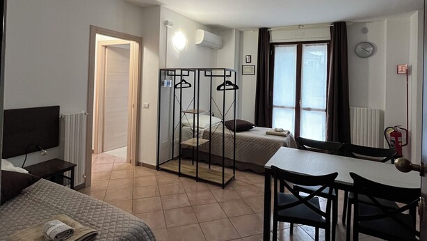 A hotel room with a bed, a desk, and a chair. There is a window with curtains, a radiator, and a clock on the wall. - Quadrifoglio affittacamere (Novi Ligure)