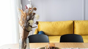 A dining table with a vase of dried flowers, a tray with glasses, and a yellow sofa in the background.