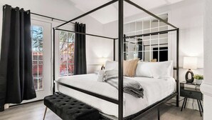A four-poster bed with a white bedspread and pillows, a black bench with gold legs, and a small bedside table with a lamp. - 5BR Luxury Oasis w/ Pool & Spa + Games and MORE! (North Las Vegas)