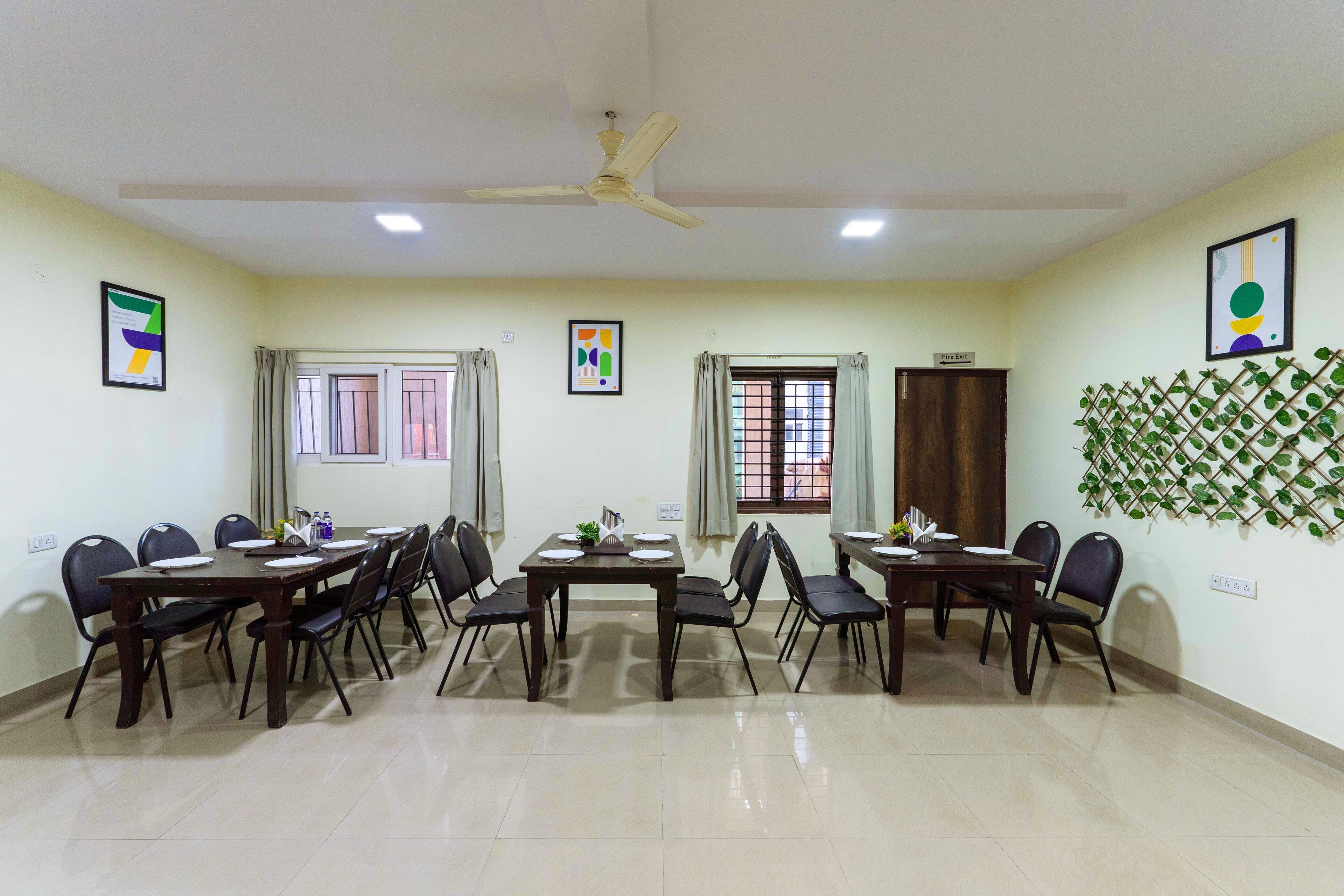 A conference room with a high ceiling, ceiling fan, and two windows with curtains. There are two tables with chairs, and a decorative plant on one table.