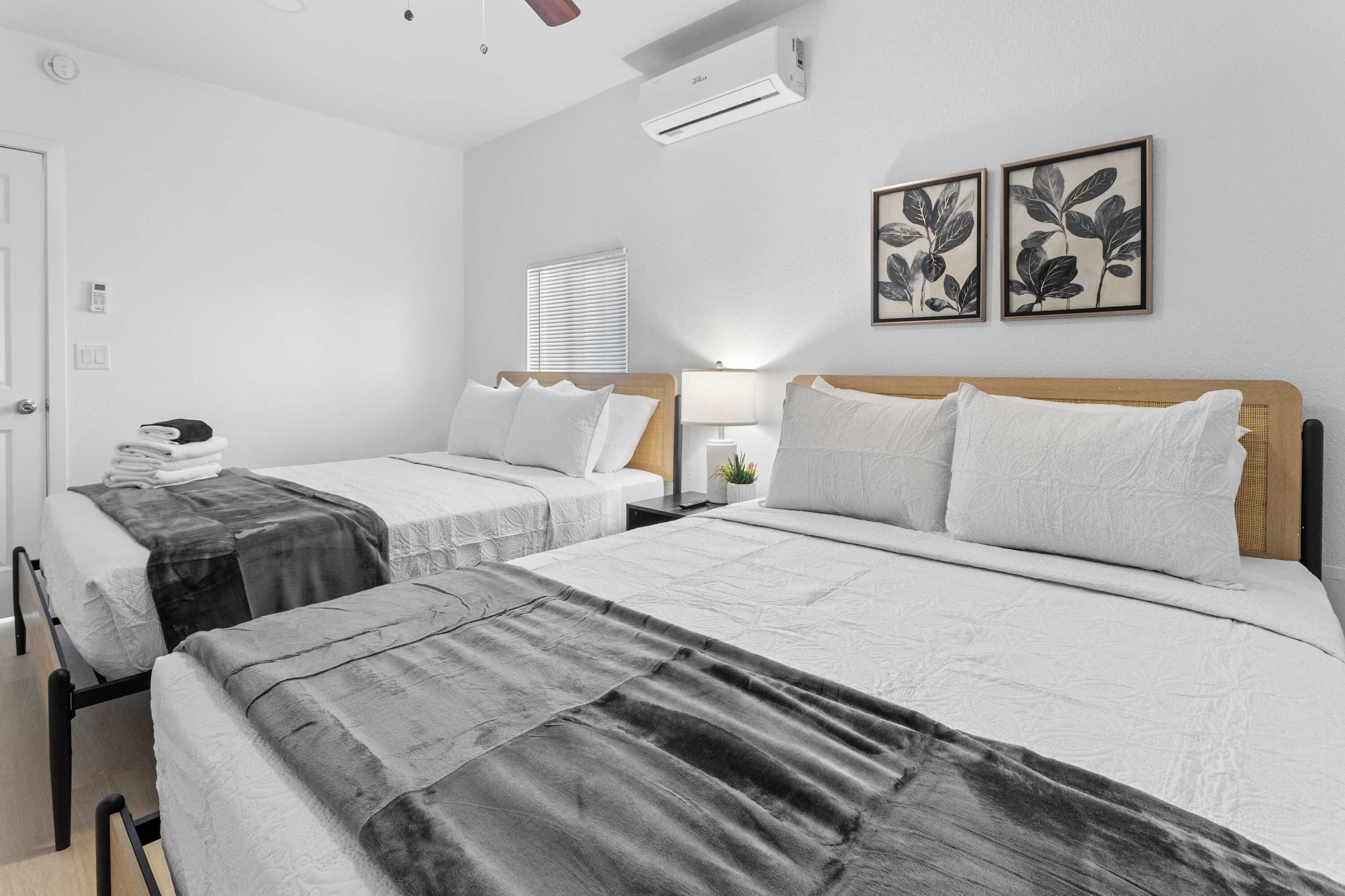 A hotel room with two beds, a bedside table with a lamp, a wall-mounted air conditioner, and two framed botanical prints.