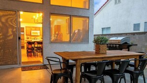 Un patio al aire libre con una mesa de madera y sillas negras, una parrilla de barbacoa y una cocina visible a través de una puerta corrediza.