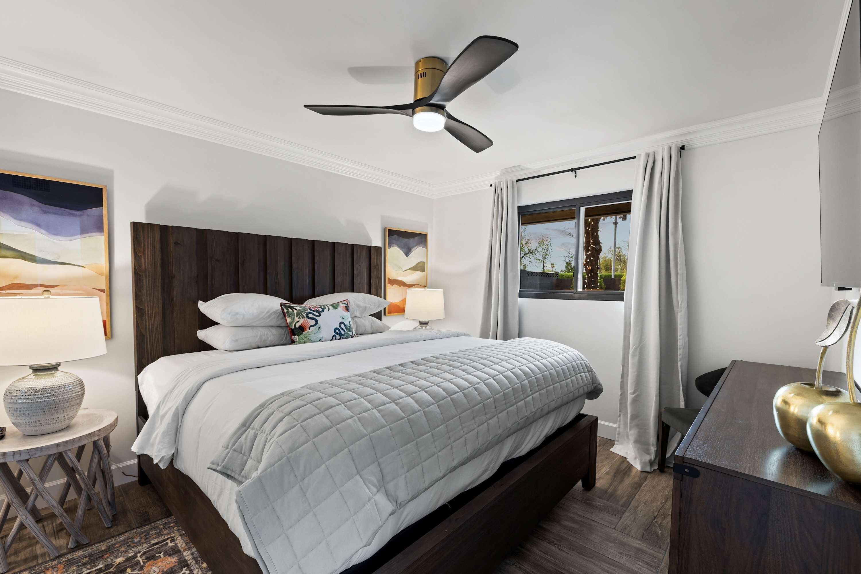 A modern bedroom with a bed, bedside lamps, a ceiling fan, and a view of the outdoors through a window.