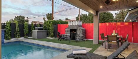 A backyard with a swimming pool, a brick outdoor fireplace, and lounge chairs.