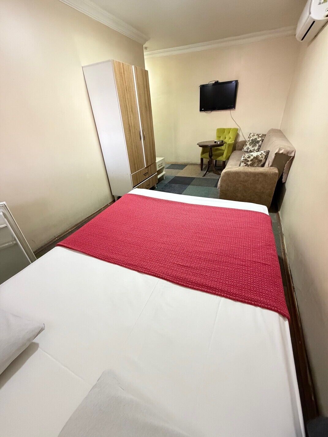 A hotel room with a bed, a wardrobe, a desk with a chair, a television, and an air conditioning unit.