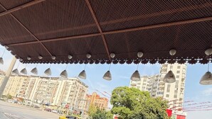 Un espacio al aire libre con una zona de estar cubierta, una planta en maceta y una calle concurrida, con coches y edificios al fondo.