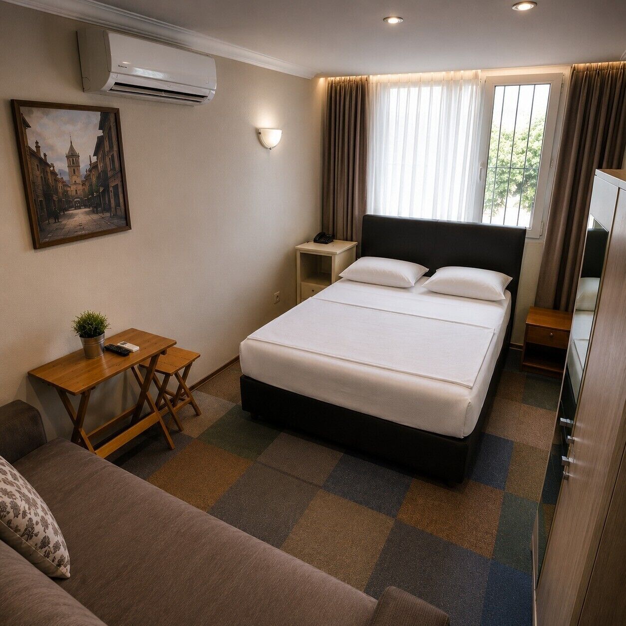 A hotel room with a bed, a desk, a chair, a small table, a plant, a painting, and an air conditioning unit.