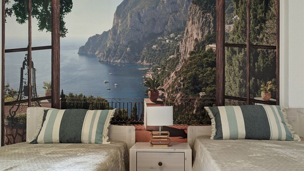 A bedroom with two beds, a nightstand, and a view of a coastal landscape through large windows. - GRAN SOL sea front apartment (Calpe)