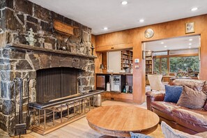 Une cheminée en pierre avec un banc, une table basse ronde en bois, un canapé en cuir et une bibliothèque dans une pièce aux murs en bois et dotée d’une grande fenêtre.