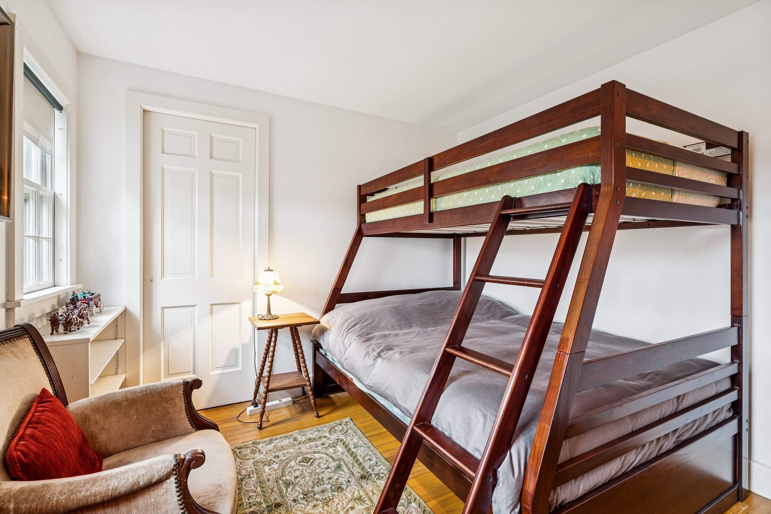 A bunk bed with a wooden frame, a chair with a red cushion, a small table with a lamp, and a door.