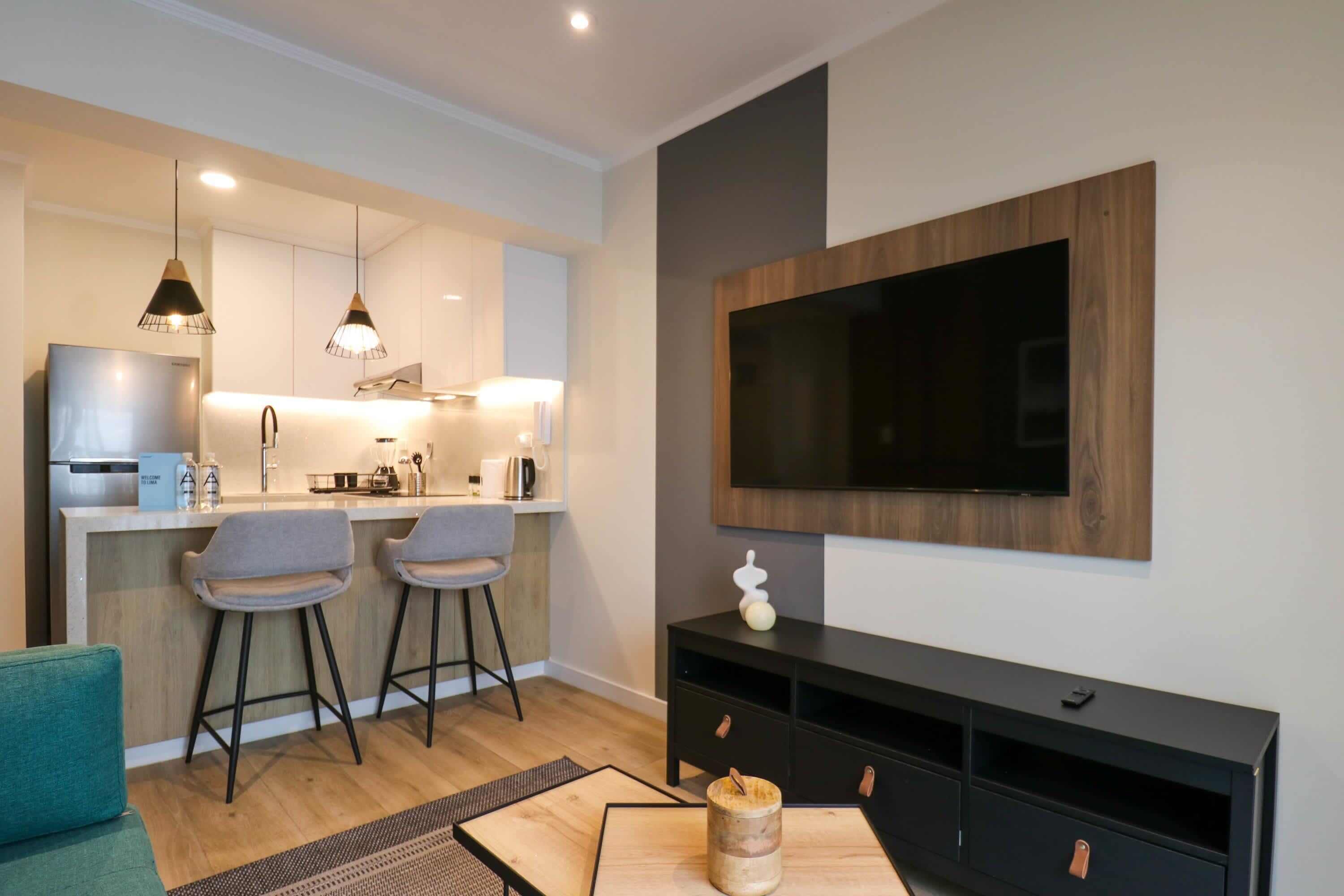 A modern living room with a flat-screen TV mounted on the wall, a wooden coffee table, two bar stools, and a sofa.