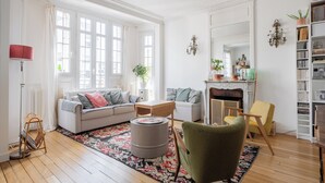 Un salon bien éclairé, avec un canapé, un fauteuil et une petite table. On y trouve une cheminée, une bibliothèque et une grande fenêtre avec des rideaux.