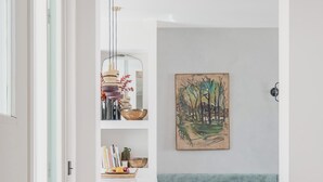 Käytävän lattia on tehty puusta kalanruotokuviossa, seinä on valkoinen ja siinä on taulu, siellä on myös puinen penkki ja kirjahylly, jossa on erilaisia esineitä.