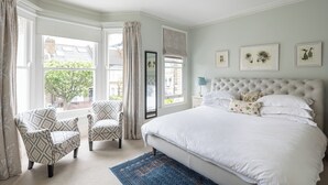 Une chambre à coucher avec un grand lit, deux fauteuils, une fenêtre avec des rideaux et des tableaux encadrés au mur.