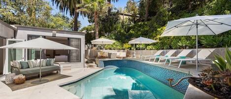 A modern outdoor pool area with lounge chairs and umbrellas, surrounded by lush greenery.