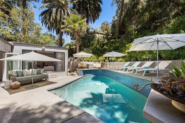 A modern outdoor pool area with lounge chairs and umbrellas, surrounded by lush greenery.