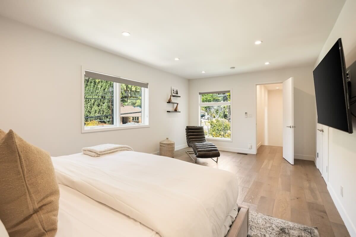 A bedroom with a large bed, a chair, a TV, and two windows offering a view of greenery.
