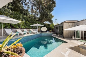 Una piscina con sillas de descanso y sombrillas, rodeada de follaje y arquitectura moderna.