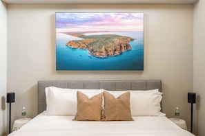 Ein Hotelzimmer mit einem Bett, Kissen, einem Nachttisch, einer Lampe und einem gerahmten Bild einer Küstenlandschaft.
