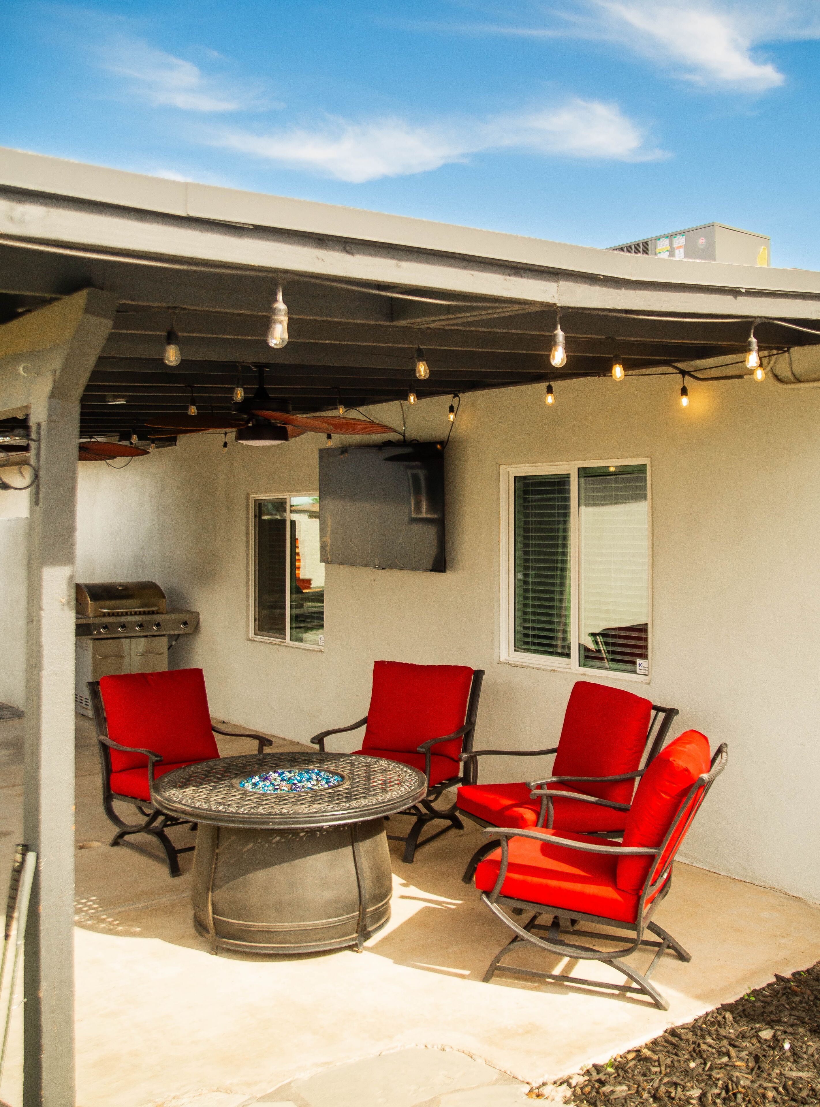 An outdoor patio with a round table and red chairs, a grill, and string lights.