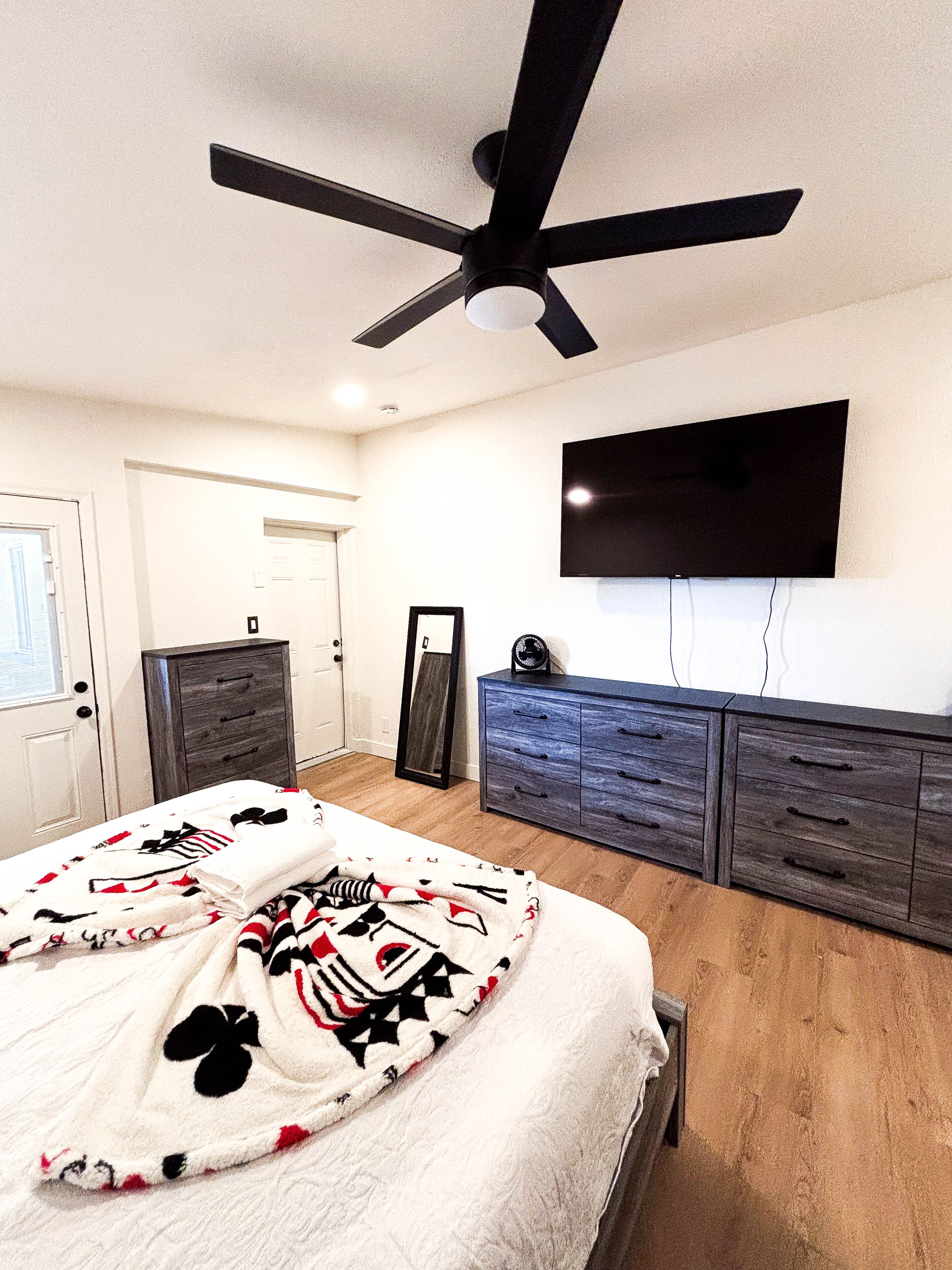 A bedroom with a bed, a ceiling fan, a dresser, a TV, and a door.
