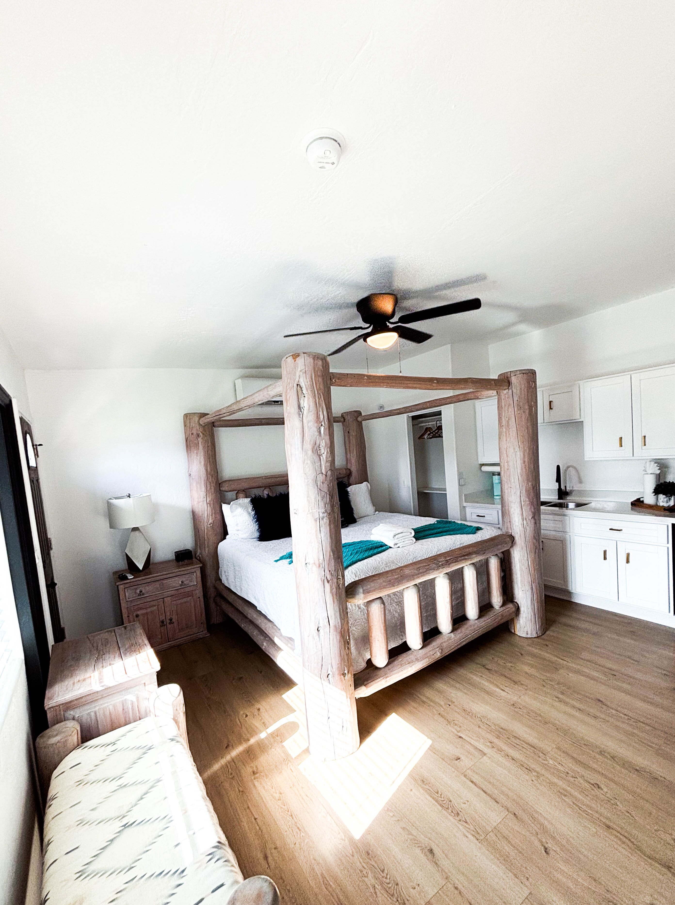 A room with a four-poster bed, a ceiling fan, a wooden floor, and white cabinetry.