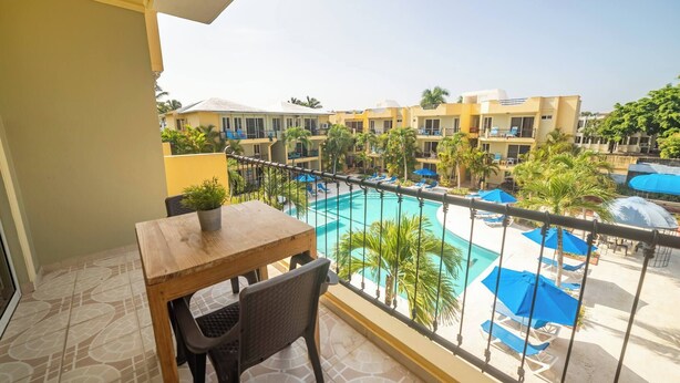 Un balcon avec une table et une chaise en bois, donnant sur une piscine bordée de chaises longues et d’ombrelles.