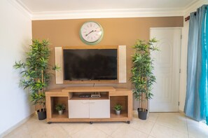 A living room with a mounted TV, a clock, a wooden entertainment unit, and two potted plants.