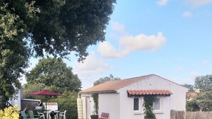 A small white house with a tiled roof, a patio with chairs and a table, and a fenced yard. - Maison de Vacances à Louer Type F2 à 10 mn des Plages, 2 Couchages 4 Personnes (Grues)
