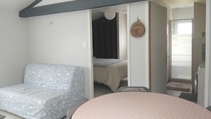 A room with a bed, a patterned sofa, a round table, and a door leading to a bathroom. - Maison de Vacances à Louer Type F2 à 10 mn des Plages, 2 Couchages 4 Personnes (Grues)