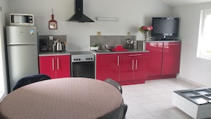 A compact kitchen with a microwave, stove, and red cabinets. A dining table is in the foreground. - Maison de Vacances à Louer Type F2 à 10 mn des Plages, 2 Couchages 4 Personnes (Grues)
