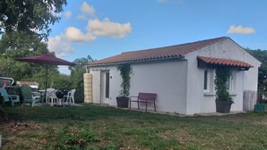 A small white building with a red-tiled roof, surrounded by a grassy yard and a few potted plants. - Maison de Vacances à Louer Type F2 à 10 mn des Plages, 2 Couchages 4 Personnes (Grues)