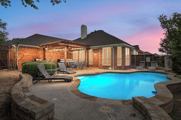 A backyard with a swimming pool, patio furniture, and a covered seating area.