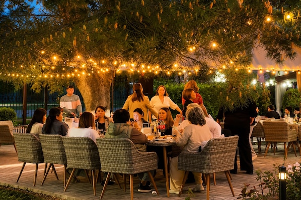 An outdoor dining area with wicker chairs and tables, string lights, and a tree in the background. - Marko Polo  (Aksay)
