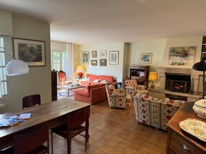 Een gezellige woonkamer met een open haard, een eethoek met een houten tafel en stoelen, en een zithoek met bloemrijk gestoffeerde fauteuils.