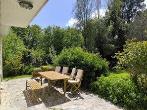Een terras buiten met een houten tafel en stoelen, omringd door weelderig groen en bomen.