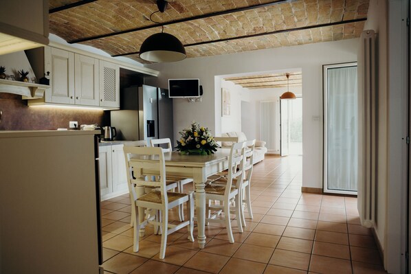 Una cocina amplia con armarios blancos, una mesa de comedor con sillas, un televisor montado sobre la encimera y un techo de ladrillo.