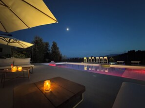 An outdoor pool area with a moon in the sky, a table with candles, and a row of chairs under a canopy.