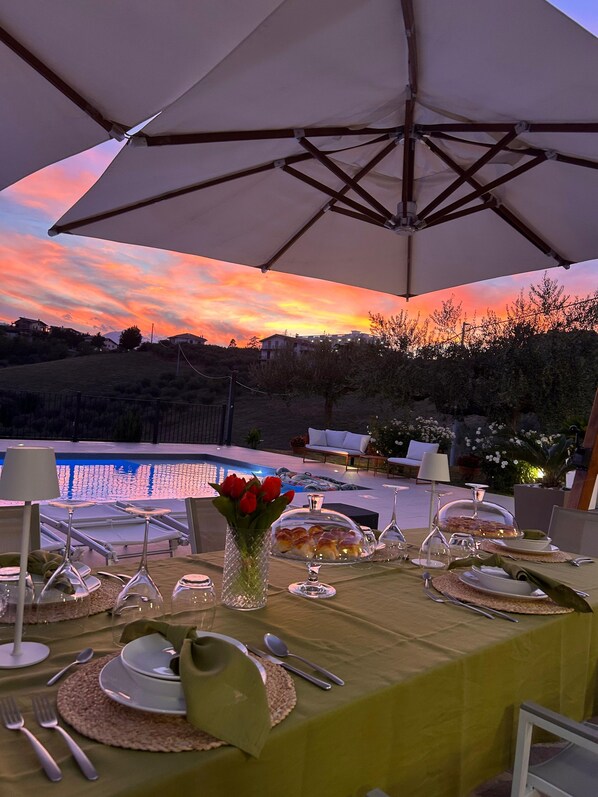An outdoor dining area with a table set for a meal, a pool in the background, and a sunset view.