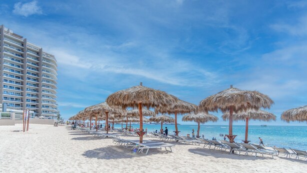 A beach with sun loungers and thatched umbrellas, a hotel building in the background, and clear blue skies.