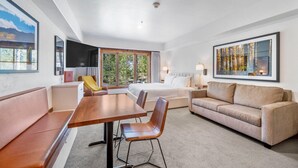 A modern hotel room with a large bed, a sofa, a dining table, and a view of greenery through the window. - Huckleberry Haven|Includes Two 1-Day Lift Tickets (Tamarack)