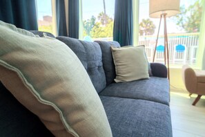 Ein graues Sofa mit beigen und weißen Kissen, eine Stehlampe und Blick auf einen Balkon mit Zaun und Bäumen.