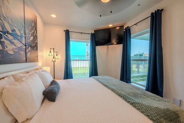 A hotel room with a bed, a flat-screen TV, a balcony with curtains, and a painting on the wall.