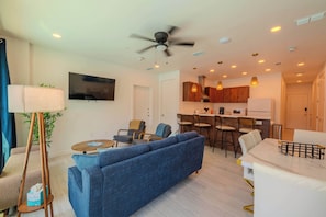 A modern living room with a blue sofa, a coffee table, and a floor lamp. There is a kitchen area with bar stools and a dining table.