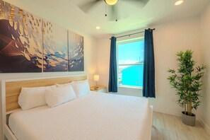 A hotel room with a large bed, a view of the sea, a potted plant, and a wall art piece.