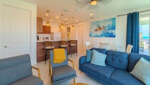 A modern living room with a blue sofa, a grey armchair, and a dining area with wooden cabinets and a white table. - Pineapple Paradise (South Padre Island)